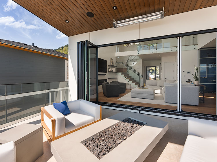 Modern patio with folding glass wall system fully opened to luxurious living room, featuring white outdoor seating and built-in fire pit